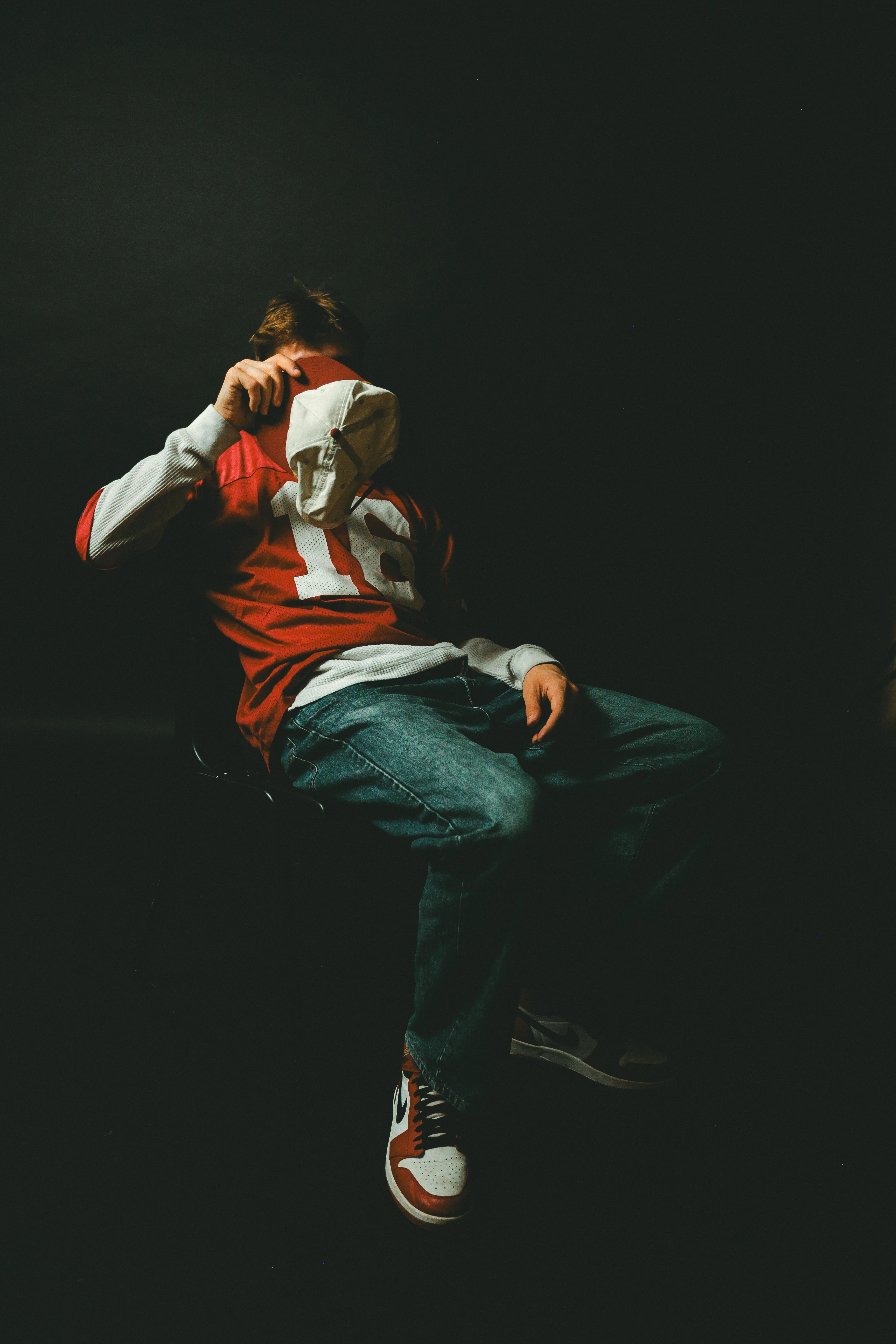 Seated figure in a red sports jersey and denim, photographed against a dark background